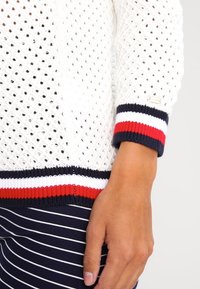 Maglione bianco lavorato a maglia con un motivo a diamante, con polsini a coste a righe blu navy, rossi e bianchi e un piccolo dettaglio con il logo.