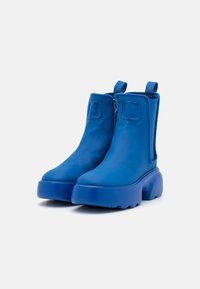 Une paire de bottes à plateforme chunky jusqu'à la cheville en bleu vif uni, avec des tirettes et des zips sur les côtés, sur un fond blanc uni.