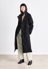Manteau trench en coton noir avec un large col, des boutons double boutonnage, des poches latérales et des poignets ajustables, associé à un jean vert clair et des bottes noires.