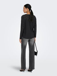 Γυναίκα με μαύρο μακρυμάνικο τοπ με τρύπες, ξεβαμμένο γκρι τζιν με ίσια μπατζάκια, μαύρα παπούτσια με τακούνι, κρίκους σκουλαρίκια και μαύρη τσάντα, κοιτάζοντας προς τα πίσω.