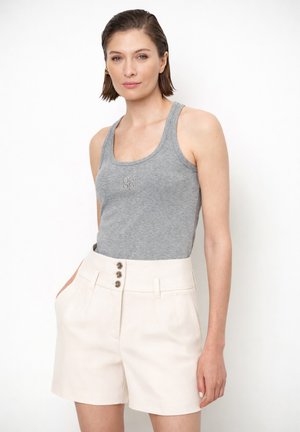 Vrouw met een grijs mouwloos topje met 0590-logo en beige shorts met hoge taille en drie knopen aan de voorkant, staand tegen een witte achtergrond.