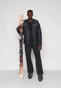 Άνδρας με μαύρο σκι κιλόνι και παντελόνι, κρατώντας ένα ζευγάρι σκι σε όρθια θέση, φορώντας μποτάκια σκι, στέκεται μπροστά σε απλό φόντο.