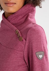 Donna che indossa un pullover a collo alto di colore bordeaux con tre bottoni decorativi in legno sul colletto e una piccola toppa sulla manica.