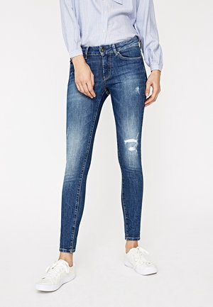 Femme portant un jean skinny bleu usé, des baskets blanches et une chemise rayée bleu clair à boutons, debout devant un fond blanc uni.