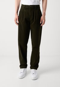 Oliwkowe spodnie z kordurowej tkaniny z dwoma przodami z zakładkami, z zapięciem na guziki i kieszeniami bocznymi, w połączeniu z białymi trampkami. Gładka faktura.