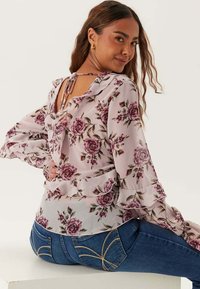 Blommig blus i ljusrosa med volangärmar och knytning i ryggen. Har ett ros-mönster och lagerdesign. Matchas med blå jeans.