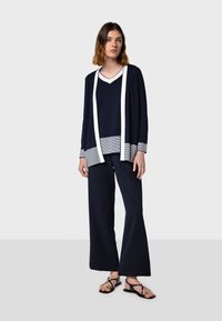 Cardigan navy con bordo bianco sopra un top a righe navy e grigio; abbinato a pantaloni wide-leg navy e sandali neri con accenti dorati.