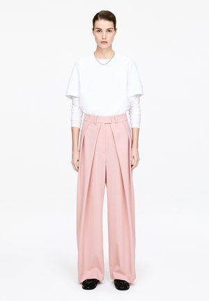 Kobieta ubrana w białą warstwową bluzkę z długim rękawem, różowe szerokie spodnie z wysokim stanem, czarne buty i srebrny łańcuszek, stojąca na tle białego tła.