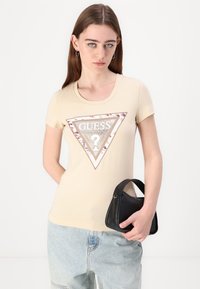 T-shirt bege de manga curta com um grande gráfico de triângulo e o logótipo "GUESS", contornado por tachas. O modelo segura uma pequena mala preta.