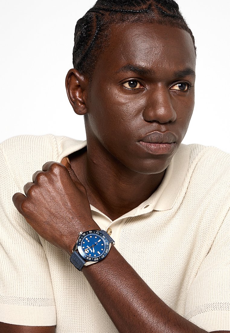 Joven con el cabello trenzado que lleva puesta una camiseta de polo beige, descansando el brazo sobre el pecho, mostrando un reloj deportivo azul en la muñeca.