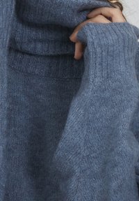 Weicher blauer Strickpullover mit langen Ärmeln, der eine Hand in der Nähe des Halses bedeckt, zeigt die strukturierte Oberfläche und die gerippte Kragen_detail.