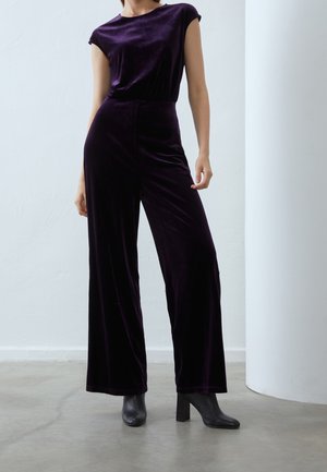 Person iført en ærmeløs mørkelilla fløjlsjumpsuit med vide ben og sorte ankelstøvler med hæl, stående på en betongulv.