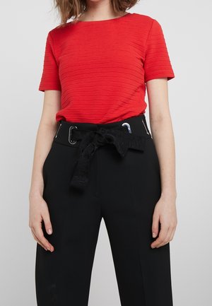 Kobieta ubrana w czerwony teksturowany krótki top włożony do wysokich czarnych spodni z szerokim paskiem zawiązanym w kokardę w talii.