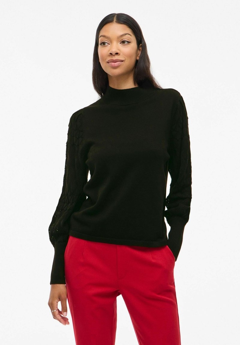 Pull noir en tricot avec un col montant et des manches texturées, associé à un pantalon rouge. Le vêtement possède des poignets ajustés et une coupe décontractée.