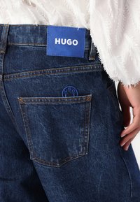 Mørkeblå denimjeans har en læraktig merkelapp med "HUGO" preget i hvitt. En sirkulær blå logo er synlig på baklommen.