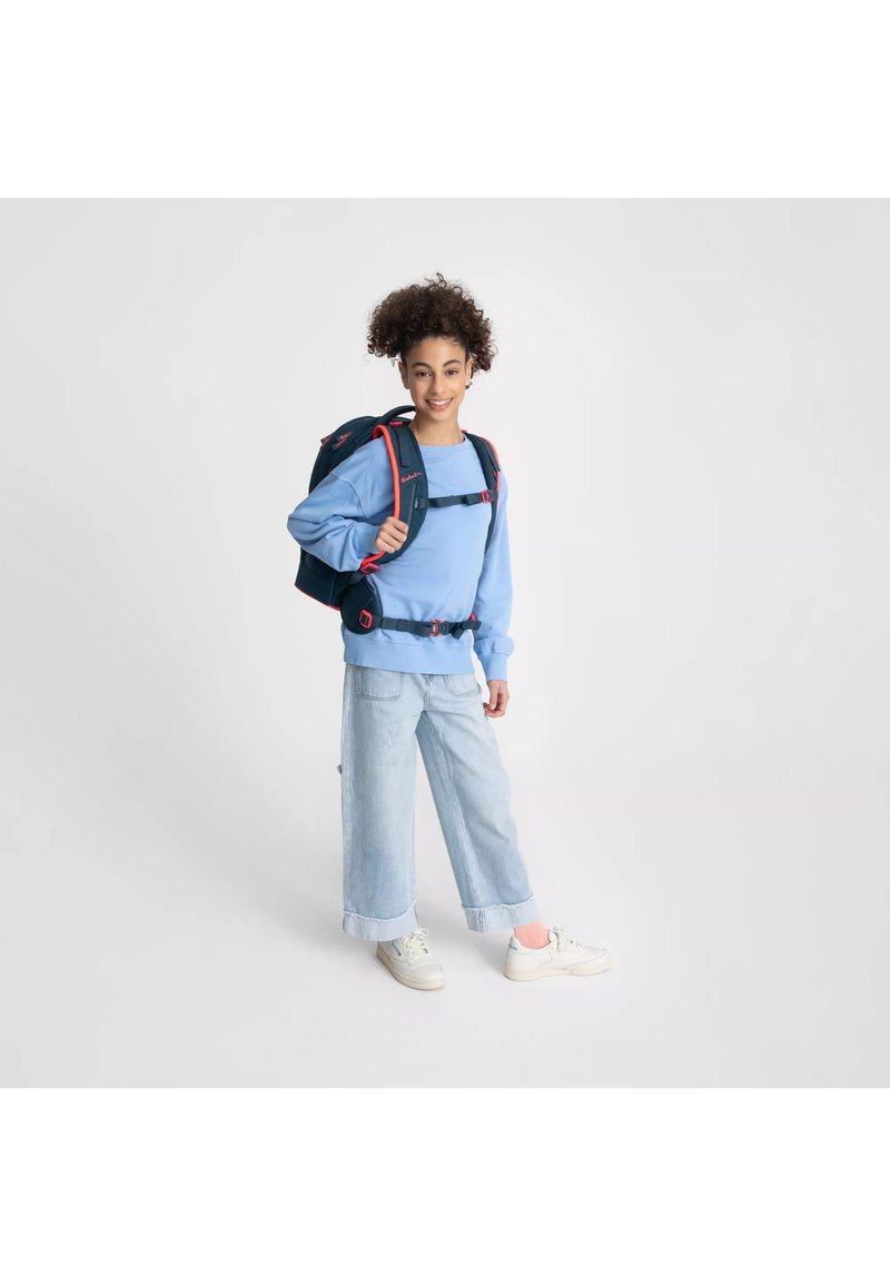 Felpa blu con una vestibilità rilassata, abbinata a jeans a gamba larga, di un lavaggio chiaro. Presenta uno zaino scuro con dettagli rossi e spallacci regolabili.