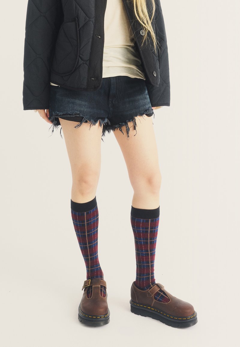 Άτομο με μαύρο καπιτονέ μπουφάν, μπεζ τοπ, ξεφτισμένα τζιν σορτς, καρό κάλτσες μέχρι το γόνατο και καφέ παπούτσια με αγκράφα, στέκεται σε φωτεινό φόντο.