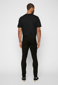 Melna pieguloša T-kreklā un melnās šaurās bikses, izgatavotas no kokvilnas maisījuma, ar aizmugurējām kabatām un redzamām šuvju detaļām.