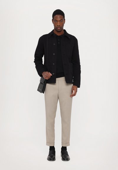 Black fabric jacket with button closure, worn over a black sweater. Light gray tailored trousers with rolled cuffs, black loafers, and a small black bag.