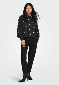 Sieviete, kas valkā melnu džemperi ar baltām margrietiņu rakstiem, melnas šauras bikses un melnas augstpapēžu potīšu botus, stāv uz viendabīgas fona.