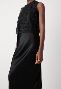 Czarny koronkowy crop top z kwiatowymi wzorami i falbaniastymi krawędziami, nałożony na gładką, satynową czarną sukienkę o zwiewnym kroju.