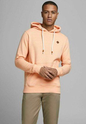 Pfirsichfarbener Hoodie mit einer Kordelzugkapuze, Fronttasche und kleinem braunem Logo-Patch auf der Brust. Weicher Stoff, lockere Passform, gerippte Manschetten.