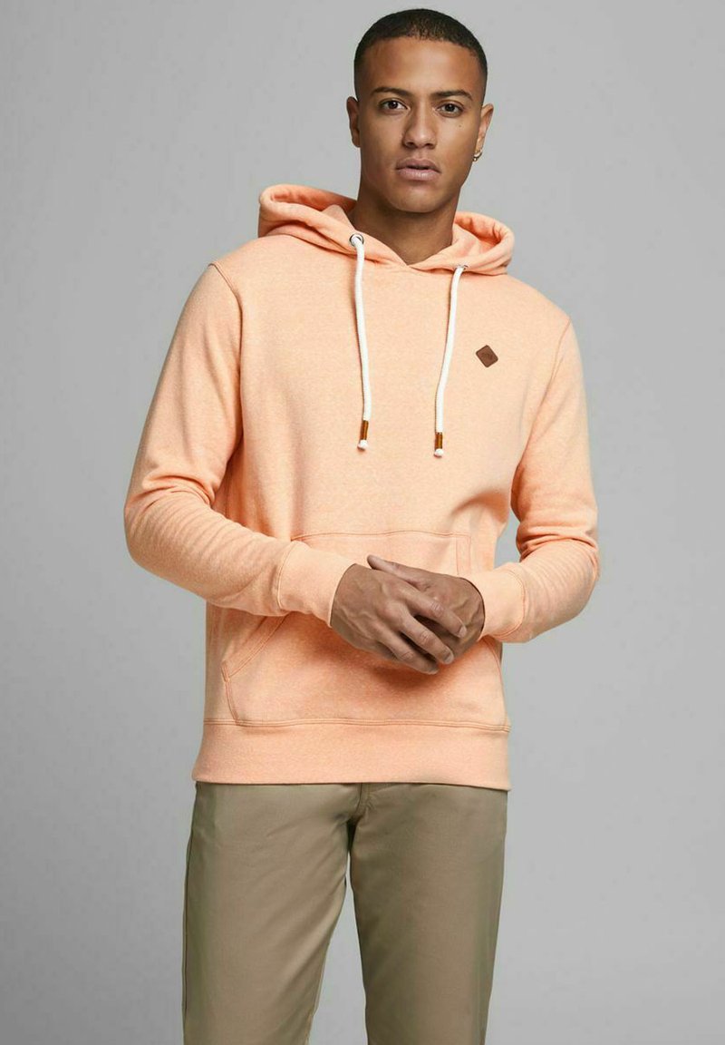 Pfirsichfarbener Hoodie mit einer Kordelzugkapuze, Fronttasche und kleinem braunem Logo-Patch auf der Brust. Weicher Stoff, lockere Passform, gerippte Manschetten.