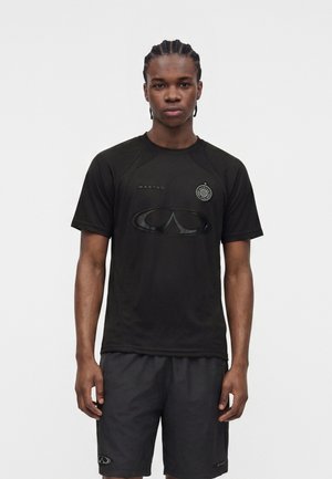 Ung mand med flettet hår iført en sort sportsskjorte med korte ærmer og sorte shorts med logoer, står mod en ensfarvet hvid baggrund.