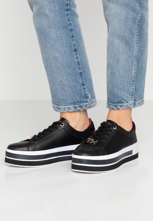 Zapatillas negras de plataforma con suela a rayas blancas y negras, logo "TH" en tono dorado en los cordones, usadas con vaqueros azul claro con puños.