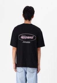 T-shirt en coton noir à manches courtes avec un texte graphique rose "Club1990 apparel" au dos. Coupe ample, col rond et design minimal.