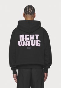 Felpa nera con cappuccio caratterizzata da un grande testo rosa sul retro che recita "NEXT WAVE" e un logo più piccolo sotto, con polsini a coste.