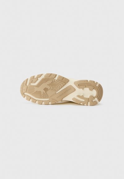 Semelle d'une chaussure en beige clair, affichant des motifs texturés et des rainures pour une meilleure adhérence. Nom de la marque "MAISON KITSUNÉ" embossé sur le côté.