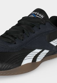 Μαύρο sneaker Reebok με λευκές λωρίδες λογότυπου, μαύρα σατινέ κορδόνια και καφέ λαστιχένια σόλα, εμφανίζεται από πλάγια και μπροστινή γωνία.