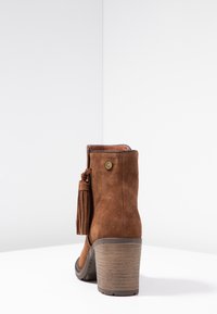 Brązowe zamszowe botki do kostki z wysokim słupkowym obcasem. Posiada szczegół z frędzlami i złoty akcent z tyłu. Teksturowana podeszwa dla lepszej przyczepności.