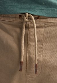 Pantalon beige avec une texture côtelée, équipé d'un cordon de serrage beige noué avec des extrémités cuivrées, associé à une chemise verte.