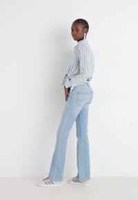 Strypet langermet skjorte knyttet rundt livet, lyseblå vide jeans og grå sneakers med tre striper. Modellen står mot en hvit bakgrunn.