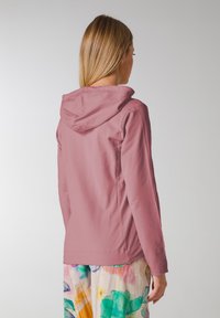 Hoodie i mjuk rosa tyg, med en avslappnad passform och långa ärmar. Modellen bär den med färgglada, mönstrade vida byxor.