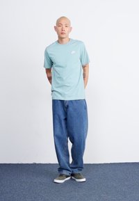 Μίντ πράσινο κοντομάνικο μπλουζάκι με λευκό λογότυπο, συνδυασμένο με φαρδιά μπλε τζιν και γκρι παπούτσια, που στέκεται πάνω σε ένα μπλε χαλί μπροστά σε λευκό τοίχο.