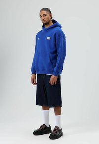 Blauwe hoodie met een voorvak, lange mouwen en een logo. Gecombineerd met donkeren denim shorts en zwarte schoenen met roze veters en witte sokken.