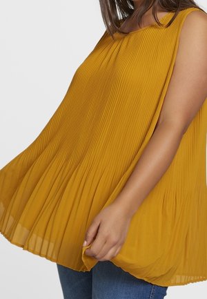 Kobieta w żółtym, plisowanym, bezrękawnym topie w kolorze musztardowym, trzymająca dół, w połączeniu z niebieskimi jeansami na jednolitym tle.