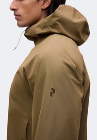 Vue de côté d'une personne portant une veste à capuche couleur fauve avec un petit logo sombre sur le haut de la manche, sur fond uni.