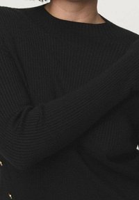 Personne portant un pull noir côtelé à manches longues avec un col rond, les bras croisés sur la poitrine.