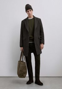 Cappotto di lana marrone sopra un maglione di lana verde, pantaloni oliva e scarpe marroni scure. Tiene una borsa tote beige e indossa un berretto abbinato.