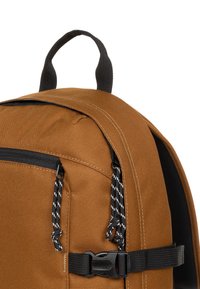 Zaino in tessuto marrone con cerniere nere, doppie tiranti a corda e una cinghia laterale con fibbia. Dotato di manico superiore per un facile trasporto.