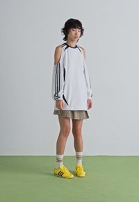 Persoon staat op een groene vloer en draagt een witte cold-shoulder lange mouwen top, een tan rok, gestreepte sokken en heldergele sneakers.