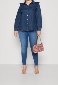 Žena v tmavé denimové blůze a modrých skinny džínách, držící růžovou texturovanou kabelku se zlatými řetízkovými popruhy, béžové otevřené boty na podpatku.