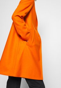 Orange uldfrakke med en afslappet pasform, der har sidelommer og synlige syningsdetaljer. Stoffet har en glat tekstur.