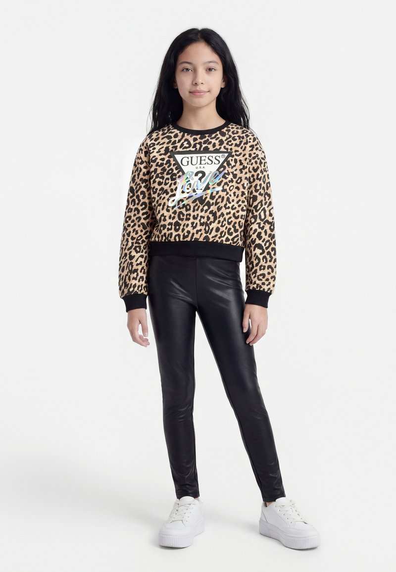 Pige iført leopardmønstret Guess-hoodie, sorte leggings i kunstlæder og hvide sneakers, stående mod en hvid baggrund.