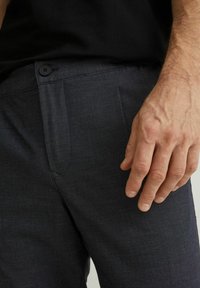 Spodnie w ciemnym kolorze z tkaniny o fakturze, zapinane na guzik w pasie, z bocznymi kieszeniami. Ręka spoczywa na nogawce spodni.