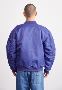 Chaqueta bomber morada con un acabado suave y brillante, puños y cuello acanalados. Cuenta con bolsillos laterales y un dobladillo ajustado.